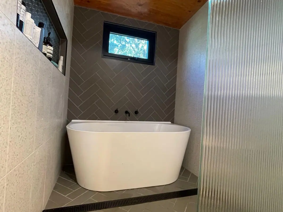 Freestanding bath against grey herringbone wall tiles under a timber-lined ceiling, with a long recessed niche and a linear floor drain, Aranda home