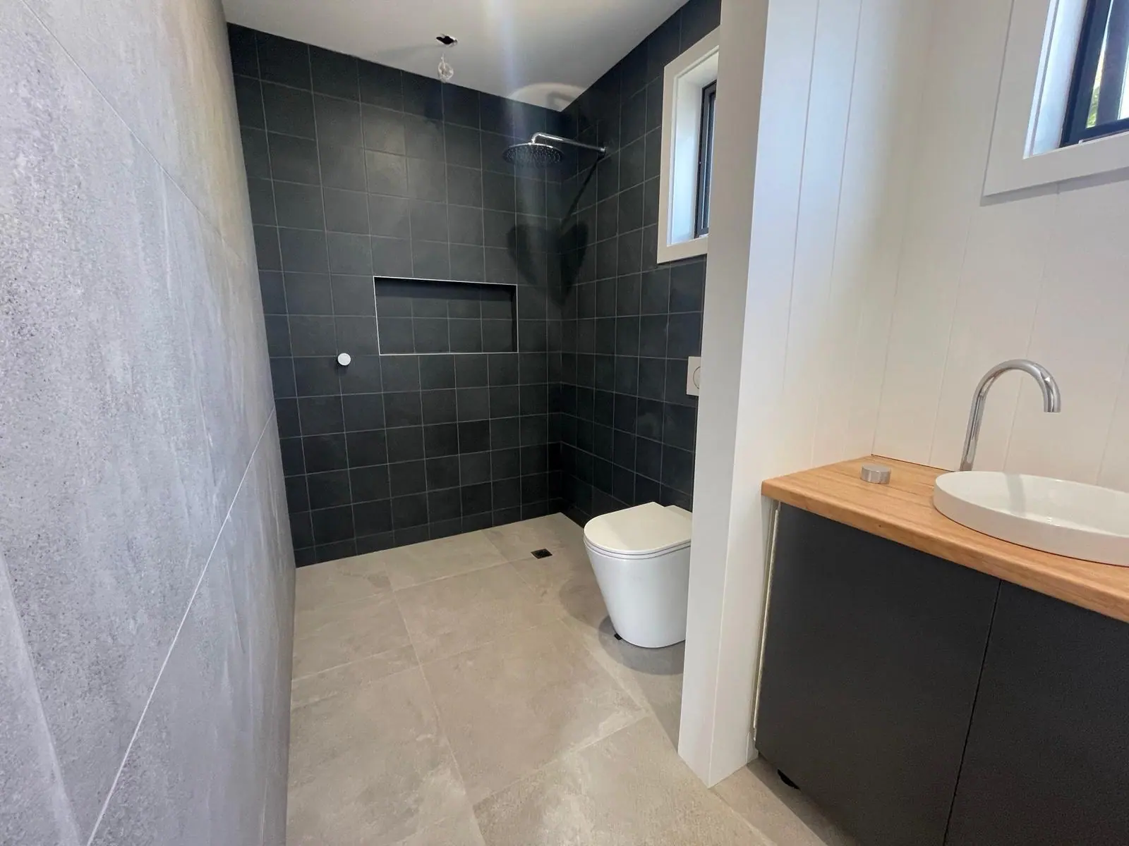 Open wet-room shower with charcoal square wall tiles and a recessed niche, a wall-faced toilet and a timber-topped vanity with a round basin, Crace home