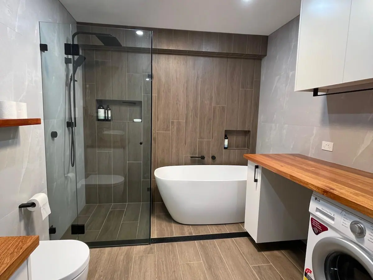 Bathroom and laundry in one room: a black-framed glass shower, a freestanding bath against timber-look wall tiles and a timber benchtop over a washing machine, Franklin home