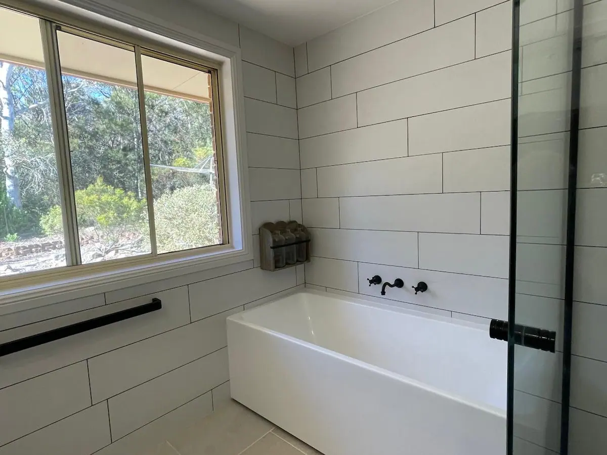 Rectangular bath beneath a window, with black wall-mounted tapware and large white wall tiles, Nicholls home