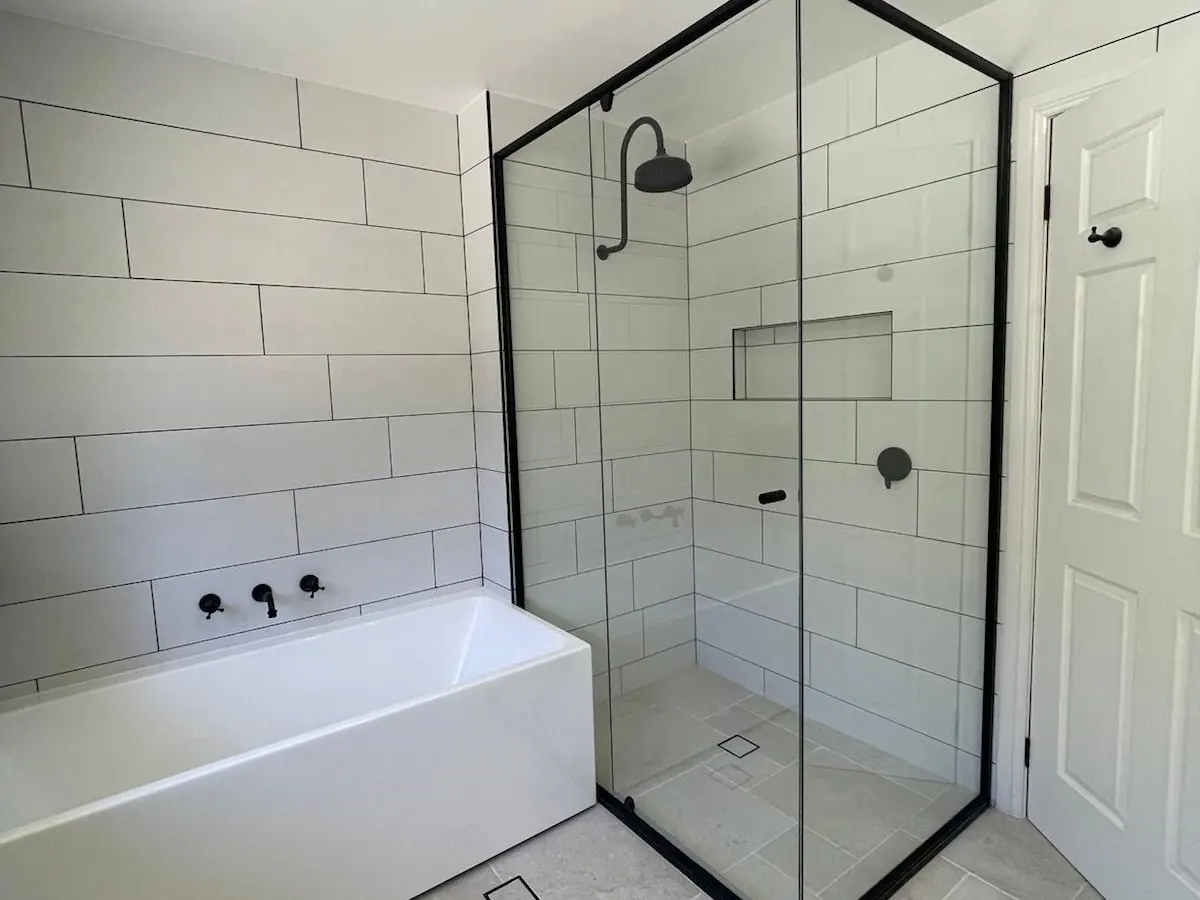 Corner shower with a black-framed glass screen and a tiled niche beside a rectangular bath, white wall tiles and stone-look floor tiles, Nicholls home