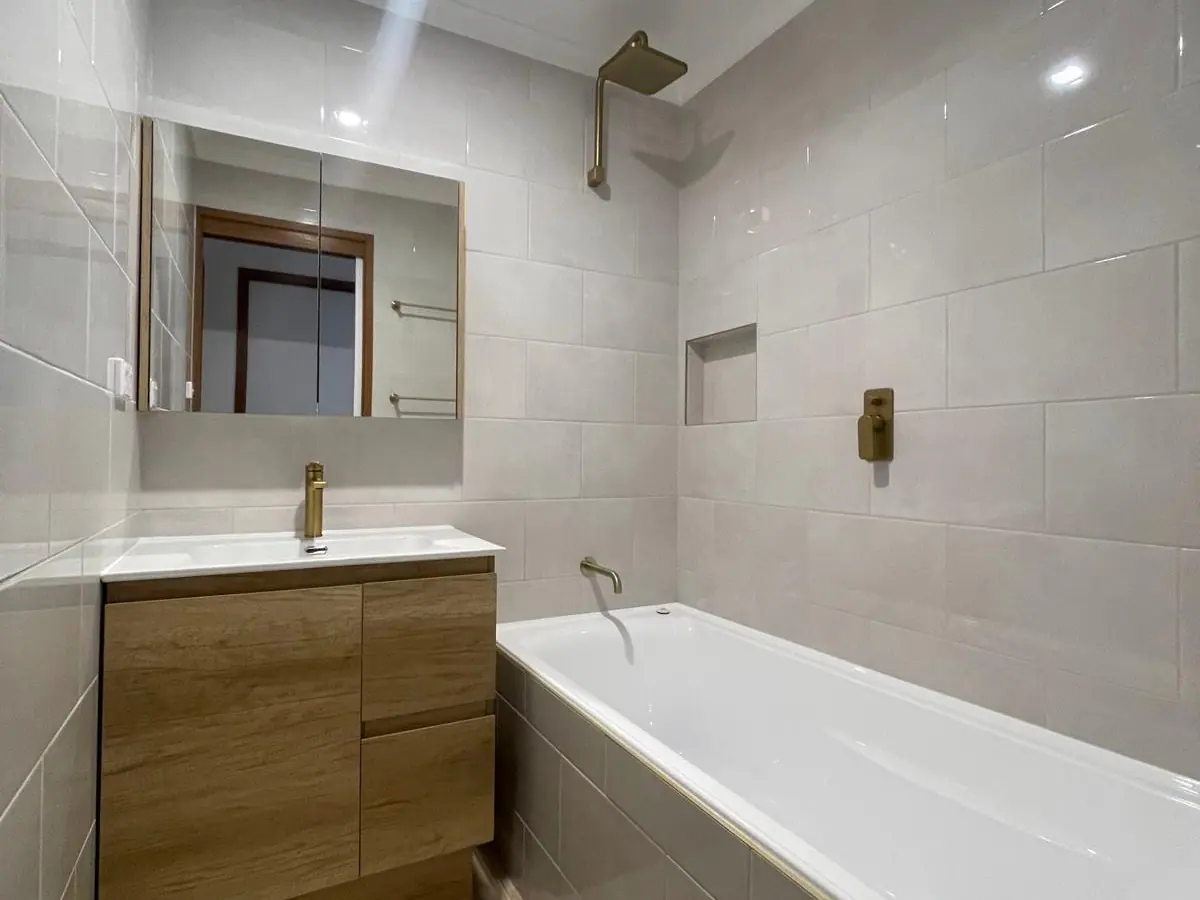 Compact bathroom with a wood-grain vanity, brushed-gold tapware, a shower over the bath and a tiled wall niche, Queanbeyan home