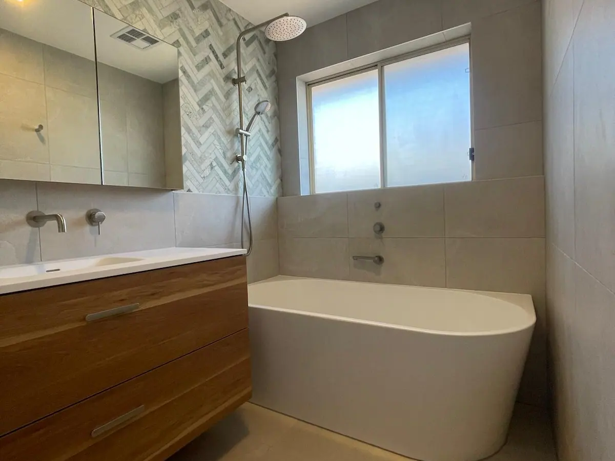 Back-to-wall bath under a window beside a green herringbone feature wall, with a timber wall-hung vanity and brushed-nickel tapware, Taylor home