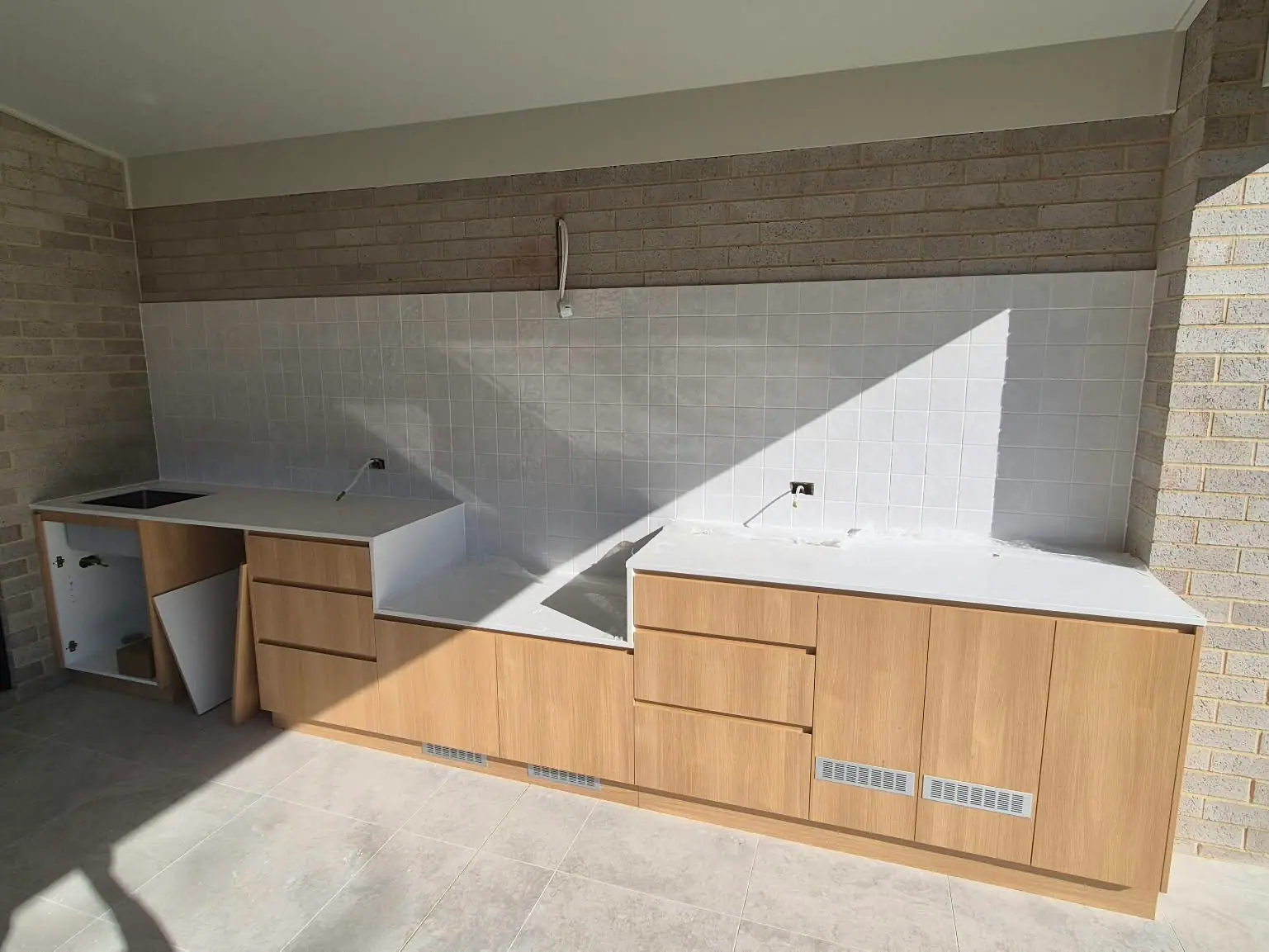 Wide view of an outdoor kitchen during installation, with timber-look cabinets and a white stone benchtop along a brick wall, Wanniassa home