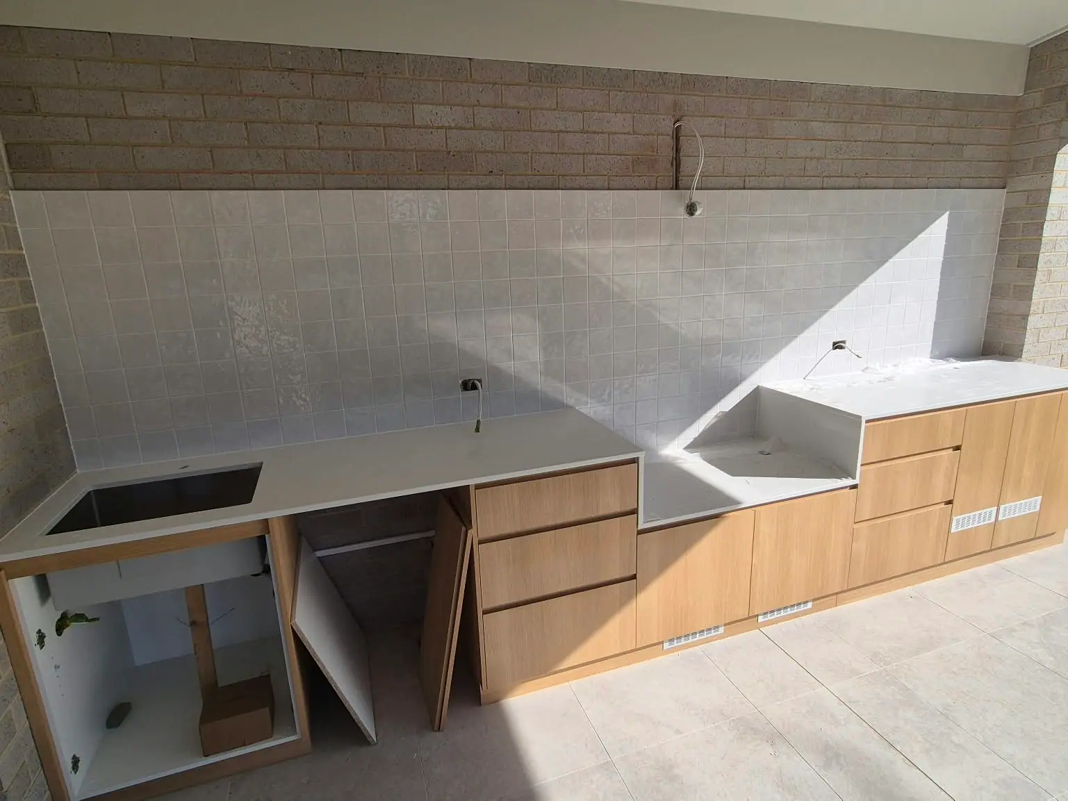 Outdoor kitchen under a covered patio during installation, with timber-look cabinets, a white stone benchtop and a white square-tile splashback, Wanniassa home