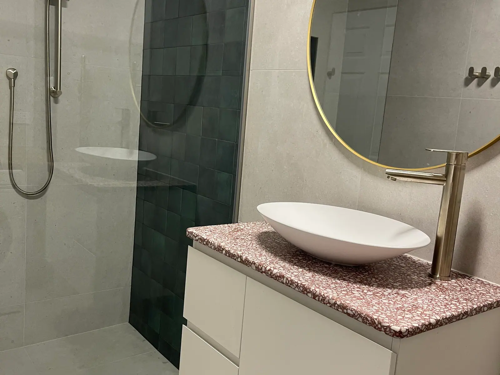 Terrazzo-topped vanity with a vessel basin, a brushed-brass tap and a round brass-framed mirror beside a walk-in shower with green wall tiles, Watson home
